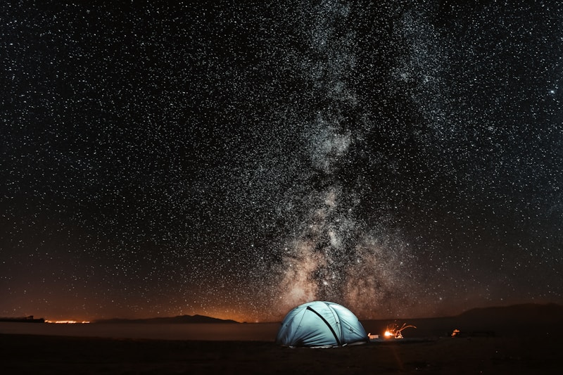Light-blue glowing dome tent on dark ground beneath a star-filled night sky with the Milky Way band visible